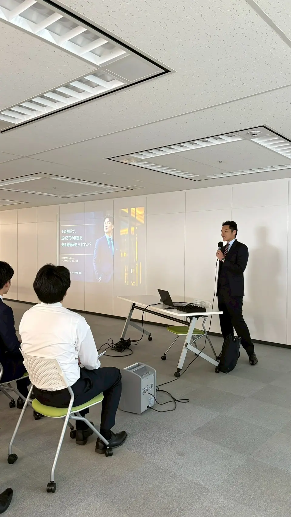 近藤が登壇し、背後の大型スクリーンに「その格好で、120万円の商品を売る覚悟がありますか?」と投影されている。受講者は後ろ姿のみ。